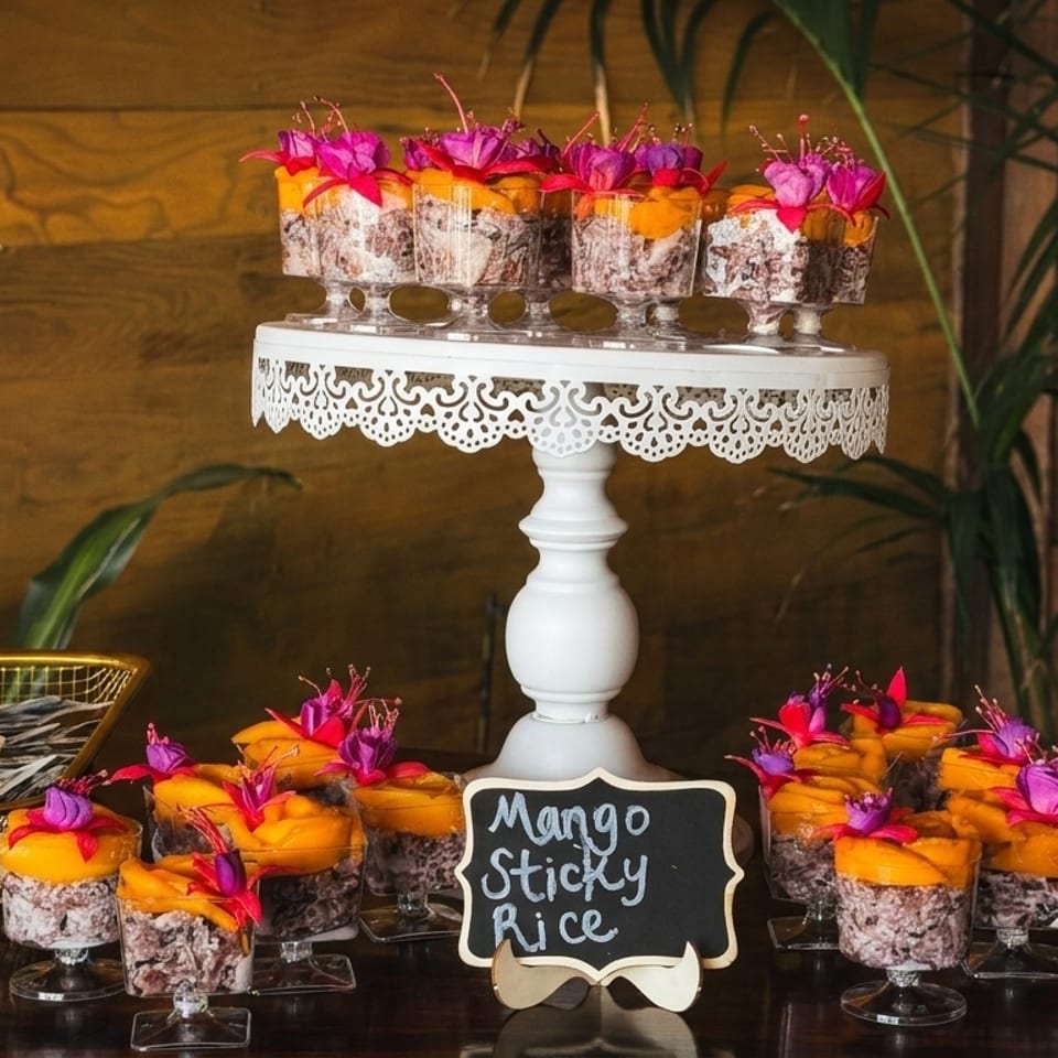 Mango sticky rice desserts in small glasses, garnished with flowers, on a white cake stand and table.