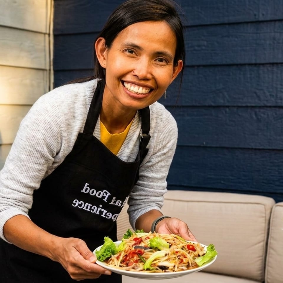 Smiling woman in an apron holding a plate of papaya salad outdoors on a patio.